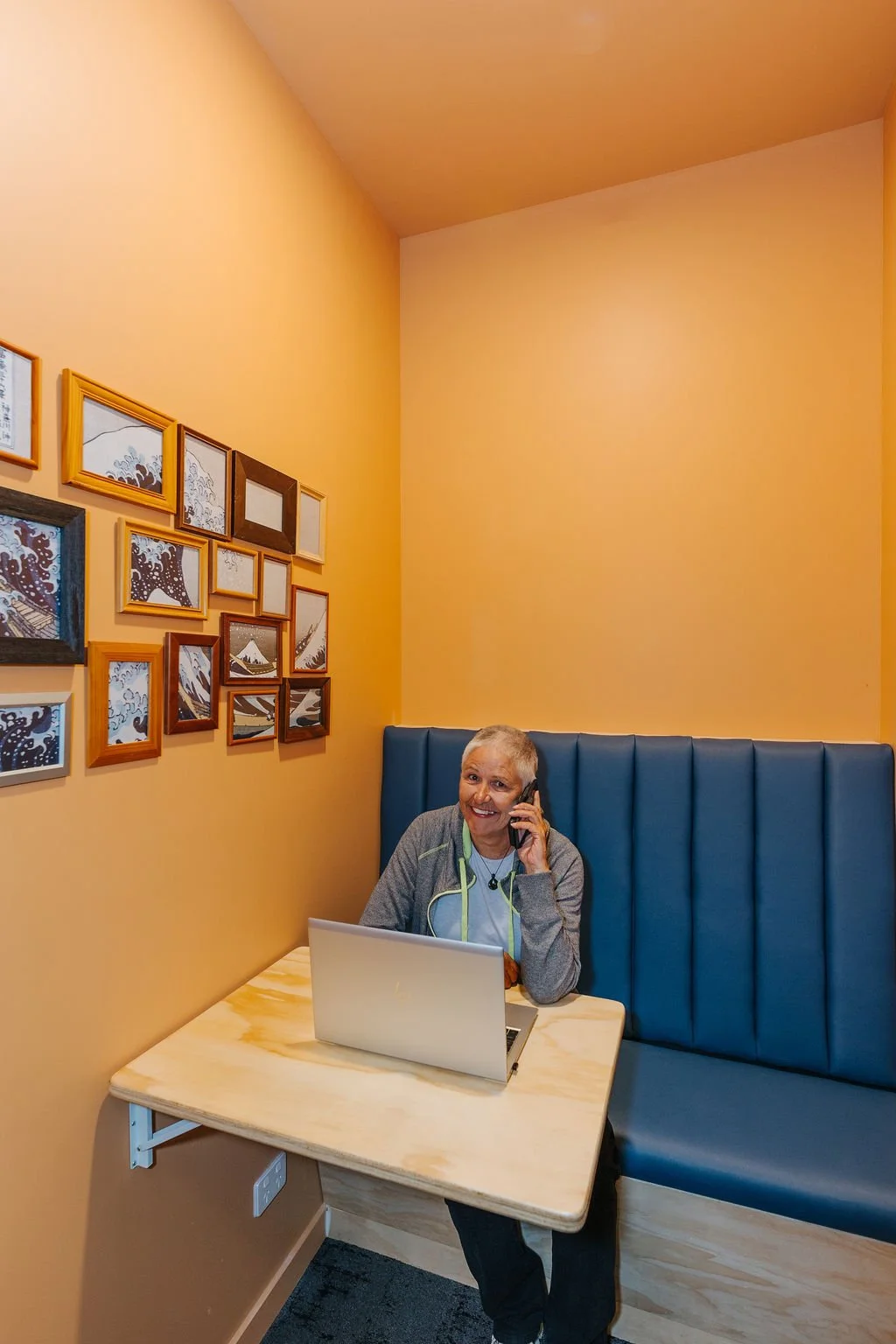 A smiling woman sitting at a small table in The Phone Booth, a little booth and waiting room in The Business Hive coworking space. She is talking on the phone with a laptop in front of her.