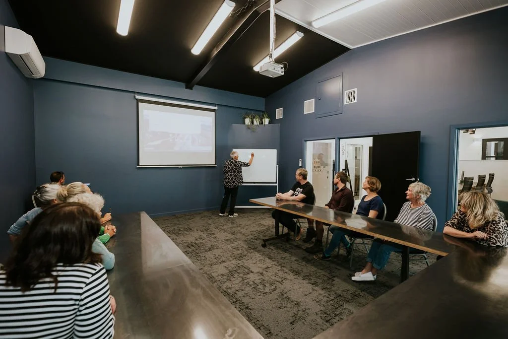 A group of people sitting at a long conference table in a room, listening to a woman who is presenting at a whiteboard and a screen.