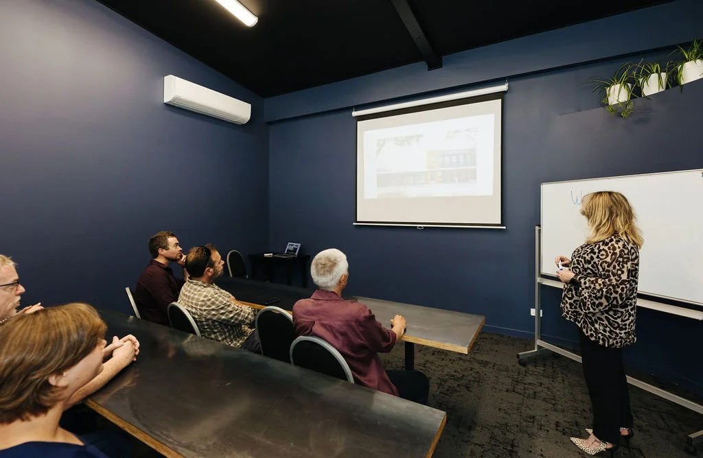 A woman is giving a presentation to a small audience in a conference room with navy blue walls. The audience members are seated at a long table, some taking notes or watching the presentation screen. A whiteboard is visible on the right side of the r