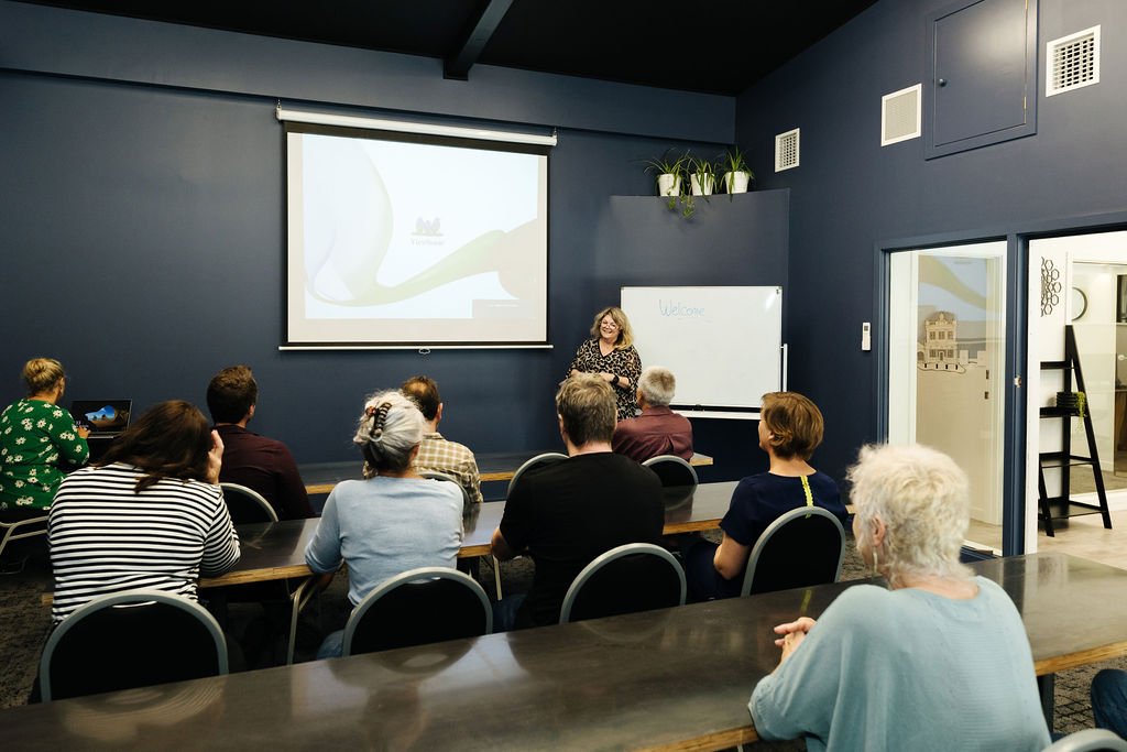 A woman is giving a presentation to a small audience in a room with dark blue walls. The audience is seated at tables, facing a whiteboard and a projection screen. There are potted plants on a shelf near the ceiling.