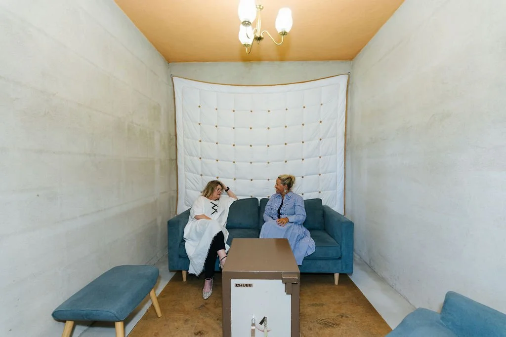 Two women sitting on a blue sofa in a small, sparsely decorated room with a white padded wall behind them and a small table in front. One woman is dressed in a white outfit with glasses, while the other wears a blue dress and has her hair in a bun.