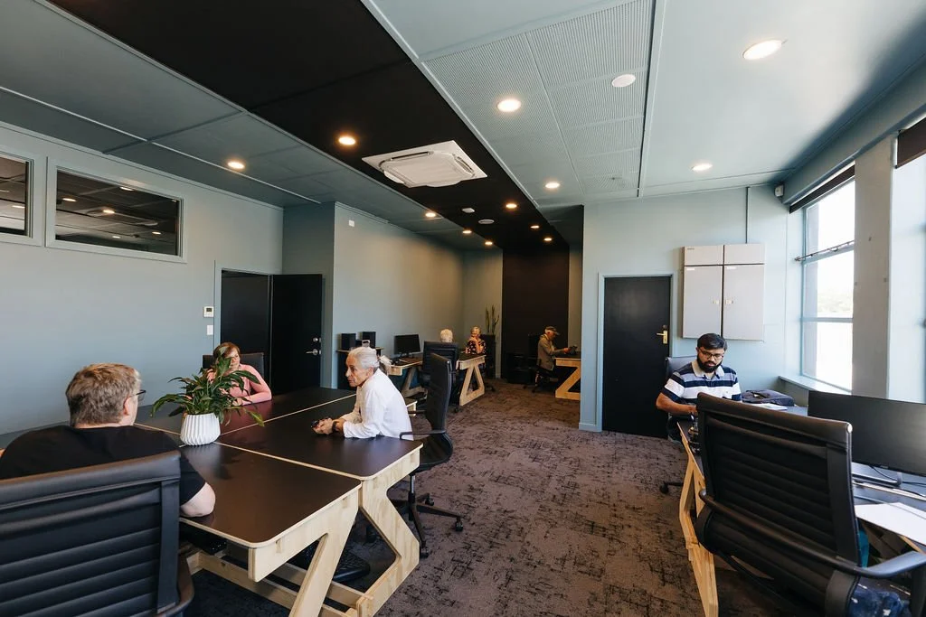 A large shared office with people working at desks and talking, with large windows on the right and a patterned carpet.