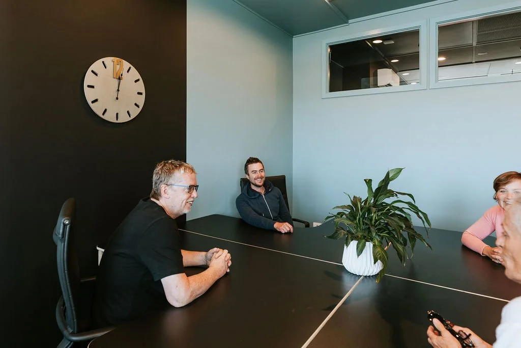 Four people in a shared office space with a black wall, a large round clock, and a potted plant on the table, smiling and talking.