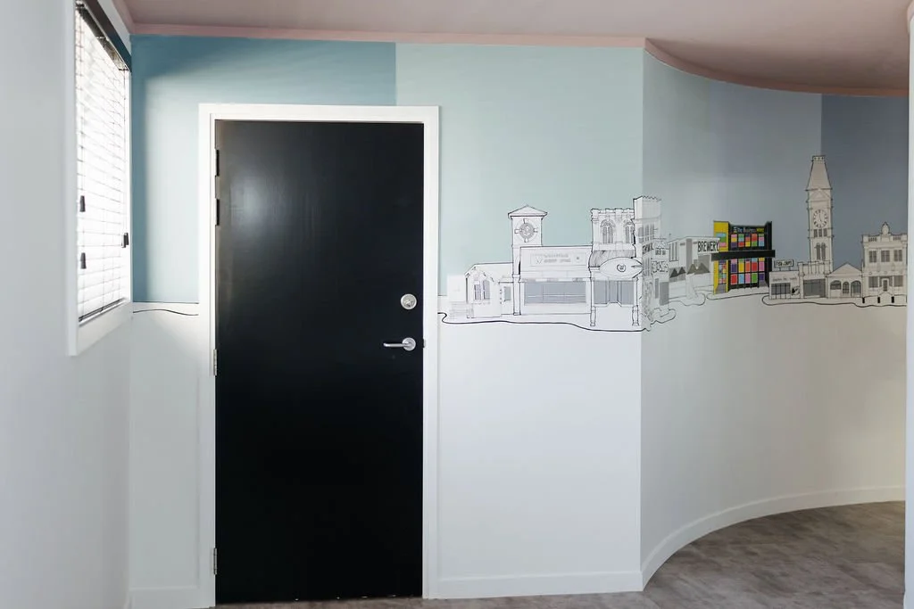 Interior room with a closed black door, light blue upper wall, white lower wall with a cityscape mural, window with blinds on the left, and gray wood flooring.