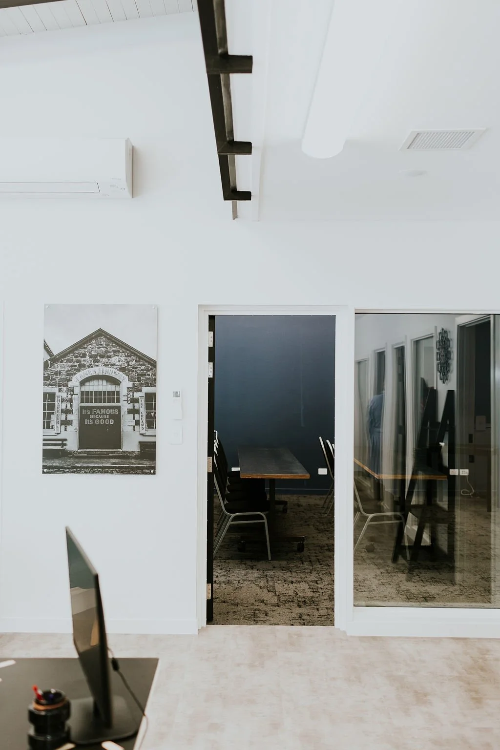 Office interior with a doorway leading to a meeting room with framed art on the wall, and a partial view of a computer monitor and desk in the foreground.