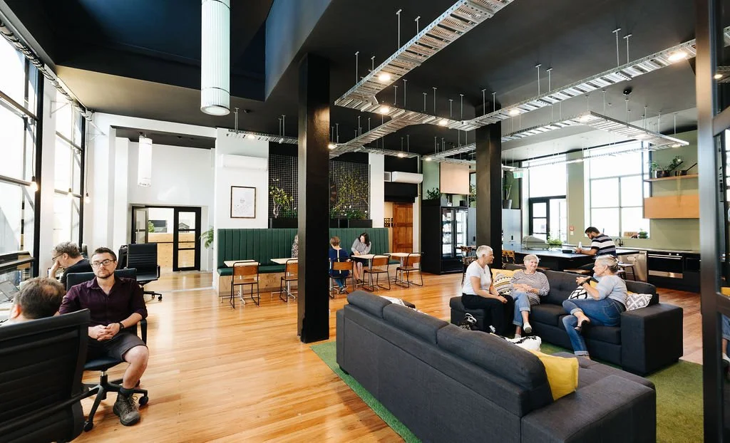Coworking space Oamaru with two people sitting on a couch and three at some tables engaging in conversation. The room is bright with white walls, a black ceiling and wooden floors