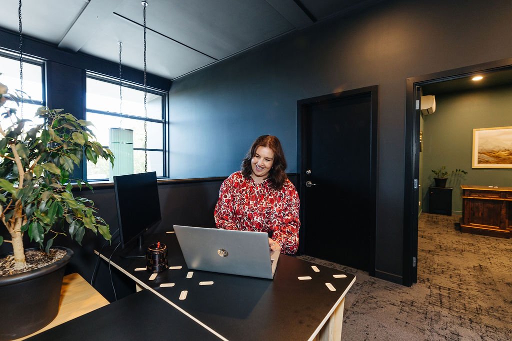 Woman smiling at a laptop sitting at a black table in a dark space light coming through the window and a door open to an office