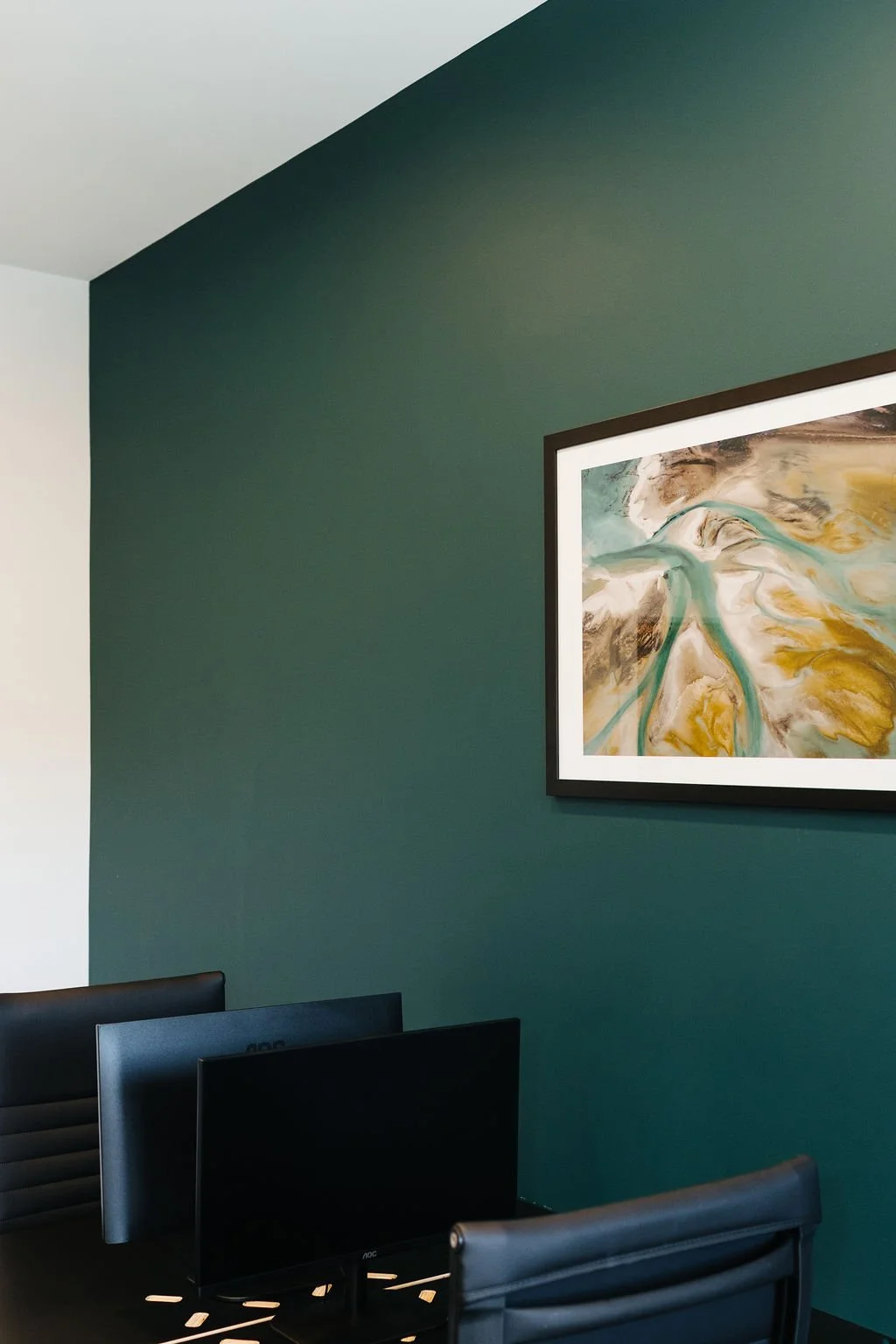 A modern leasable office meeting room with dark green accent wall, a framed abstract painting, a desk with three computer monitors, and black chairs.