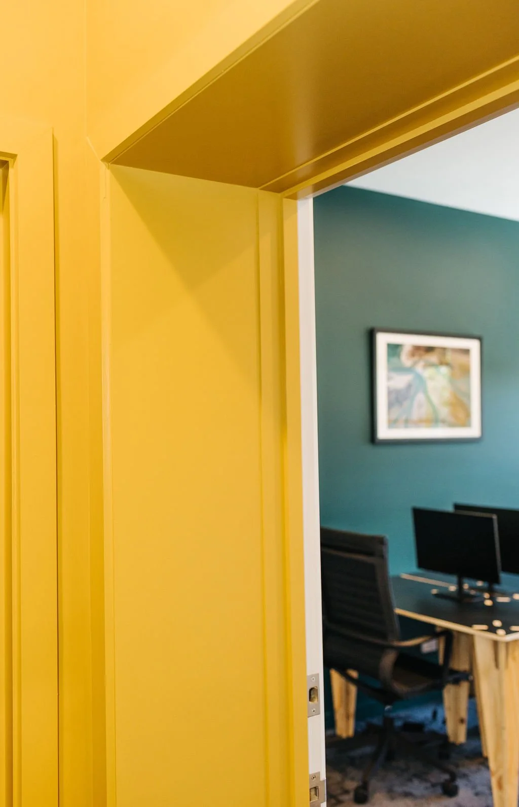 Partial view of an office with a yellow wall and door frame, and a teal-colored room with a framed artwork, desk with two monitors, and an office chair.