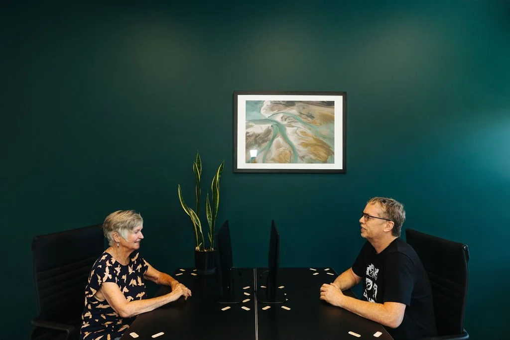 Two people sitting in a modern office at their own desks with black computer monitors, a potted plant and abstract wall art.