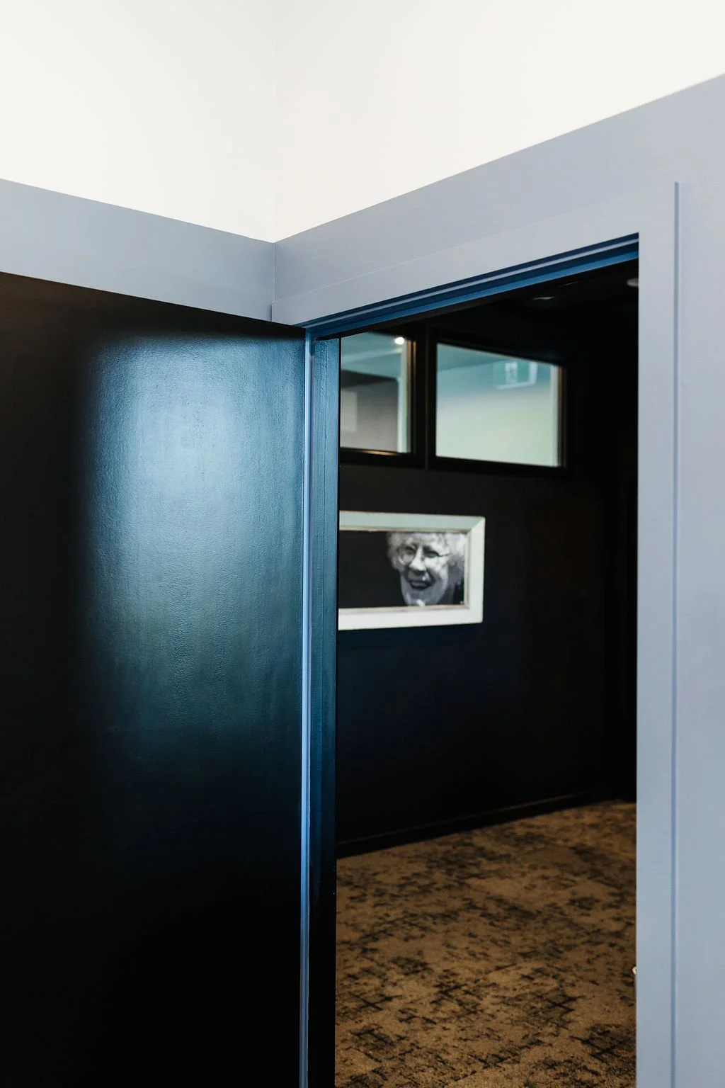 View through a doorway of an office into a coworking space with a black wall, featuring a framed black-and-white photo of an elderly woman smiling. There are small windows above the frame and a patterned carpet on the floor.