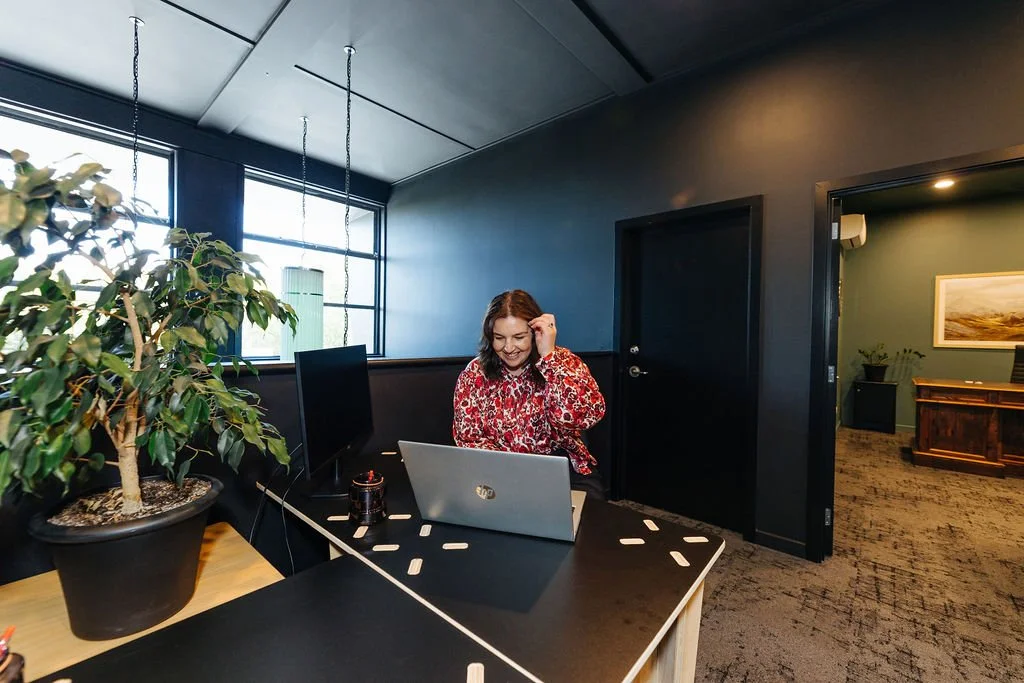 A woman sitting at a desk with a laptop, smiling and looking down, next to a large potted plant, in a modern coworking space with dark walls and large windows.