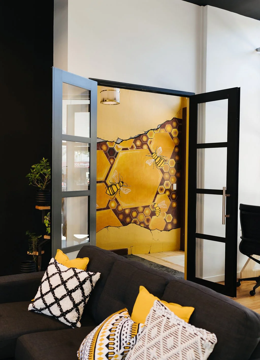 Interior of a coworking space with a black sofa, patterned pillows, potted plants, a black door open to a wall mural of bees, honeycombs, and cracked honeycomb pattern.