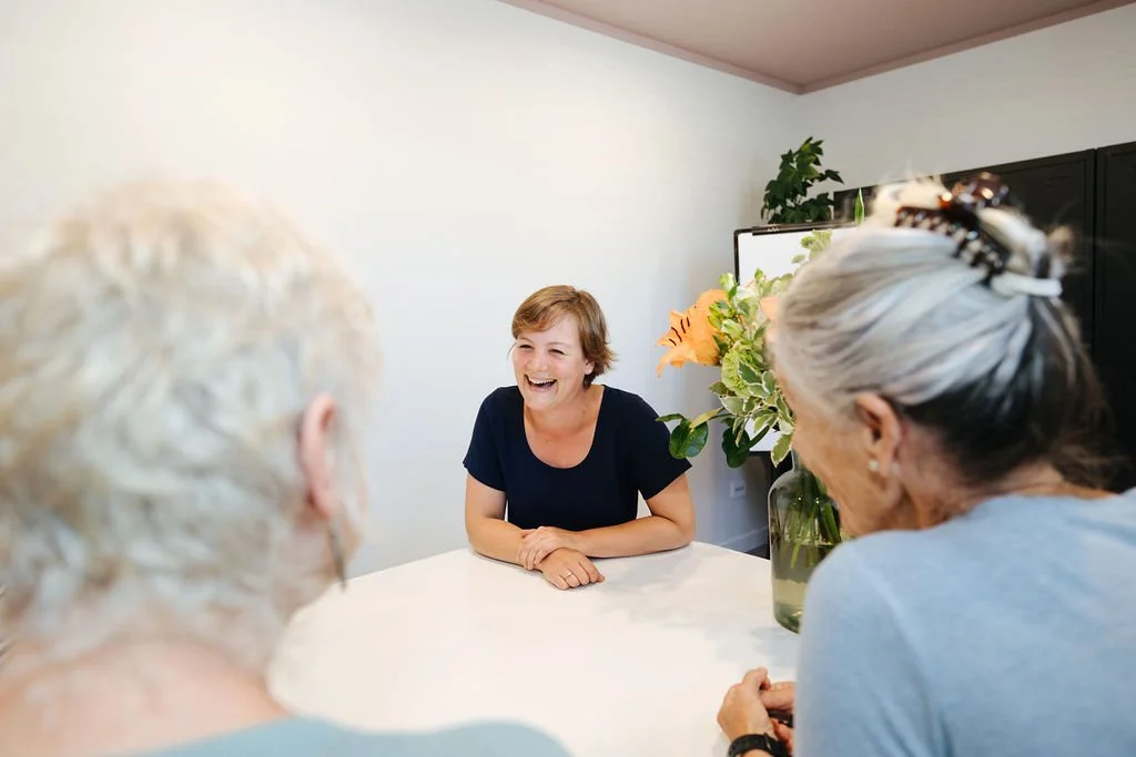 A woman sitting at a round table laughing with two older women in a meeting room with white walls and flowers on the table.
