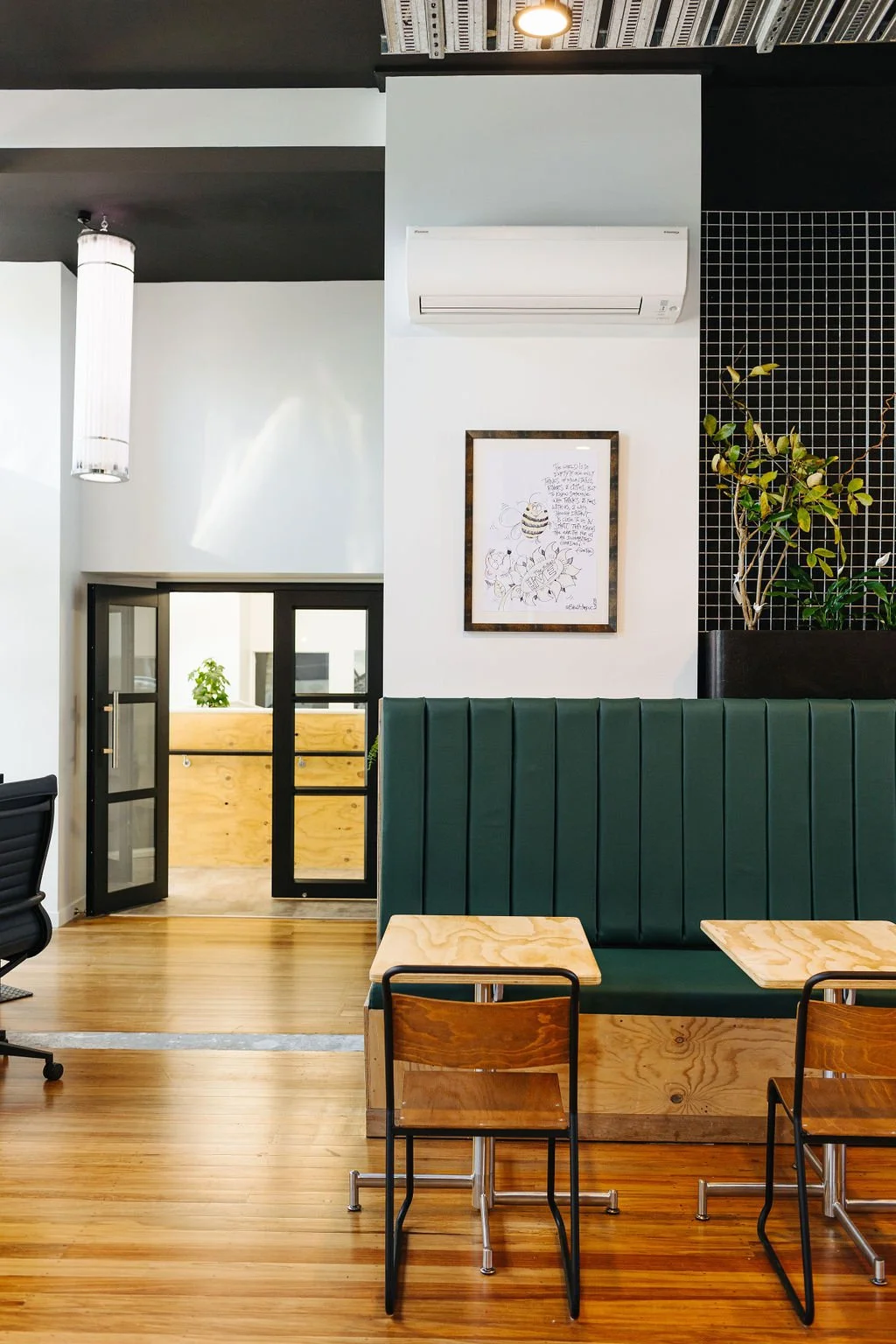 Interior of The Business Hive coworking space with wooden floors, a green upholstered booth, small wooden tables, and metal chairs. There is a framed artwork and an air conditioning unit on the white wall next to a plant, and a black grid panel. 