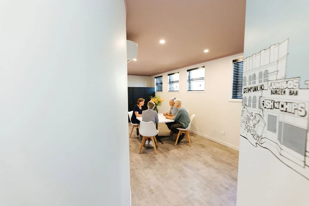 Four older women sitting around a table in a small meeting room having a conversation in a bright, modern room with beige walls, four windows with black blinds, and wooden flooring.