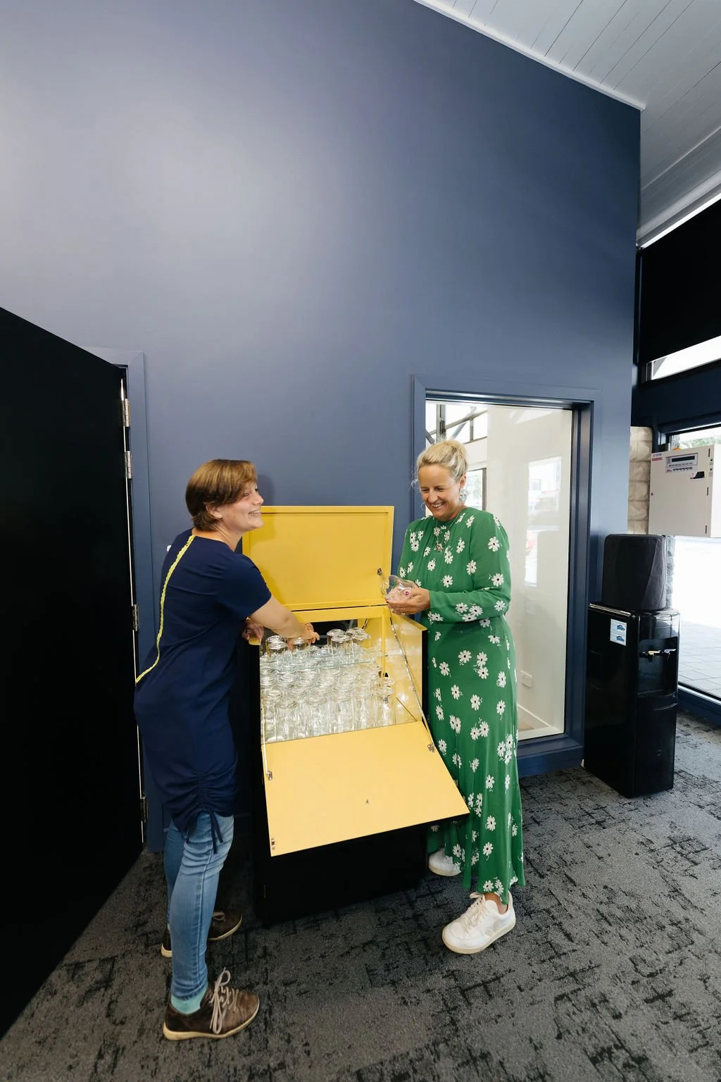 Two women smiling and talking near a yellow glassware cabinet filled with empty glasses inside a blue-walled conference room with a window and carpeted floor.
