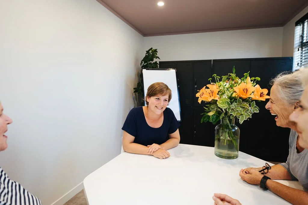 Four women sitting around a white table with a large orange flower arrangement in a glass vase, smiling in a bright meeting room.