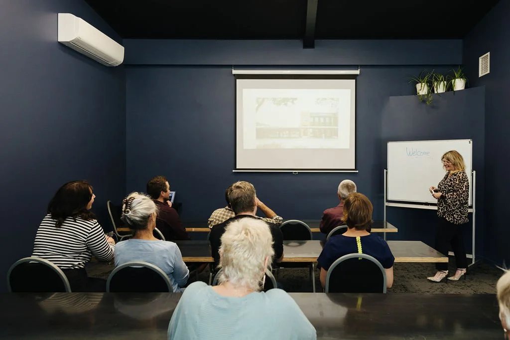 A woman standing near a whiteboard with 'Welcome' written on it speaking to a small audience in a dark blue-colored meeting room, with a presentation being projected on a screen behind her.