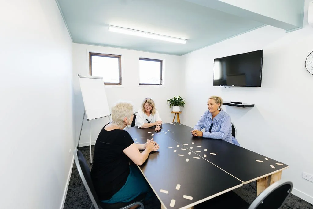 Three women sitting at a black table in a bright, white-walled meeting room. Two windows, a large flat-screen TV on the wall, and a whiteboard on a stand are visible.