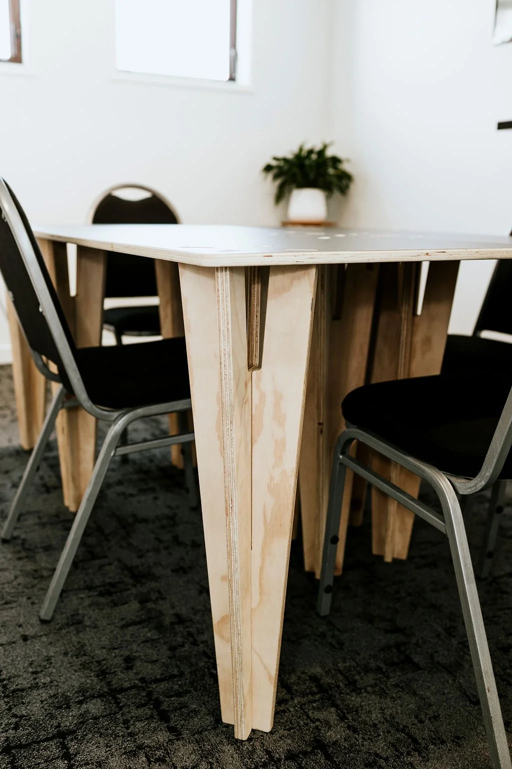 A wooden table with a unique faceted central support, surrounded by black cushioned chairs in a room with white walls and windows, and a potted plant on a shelf in the background.