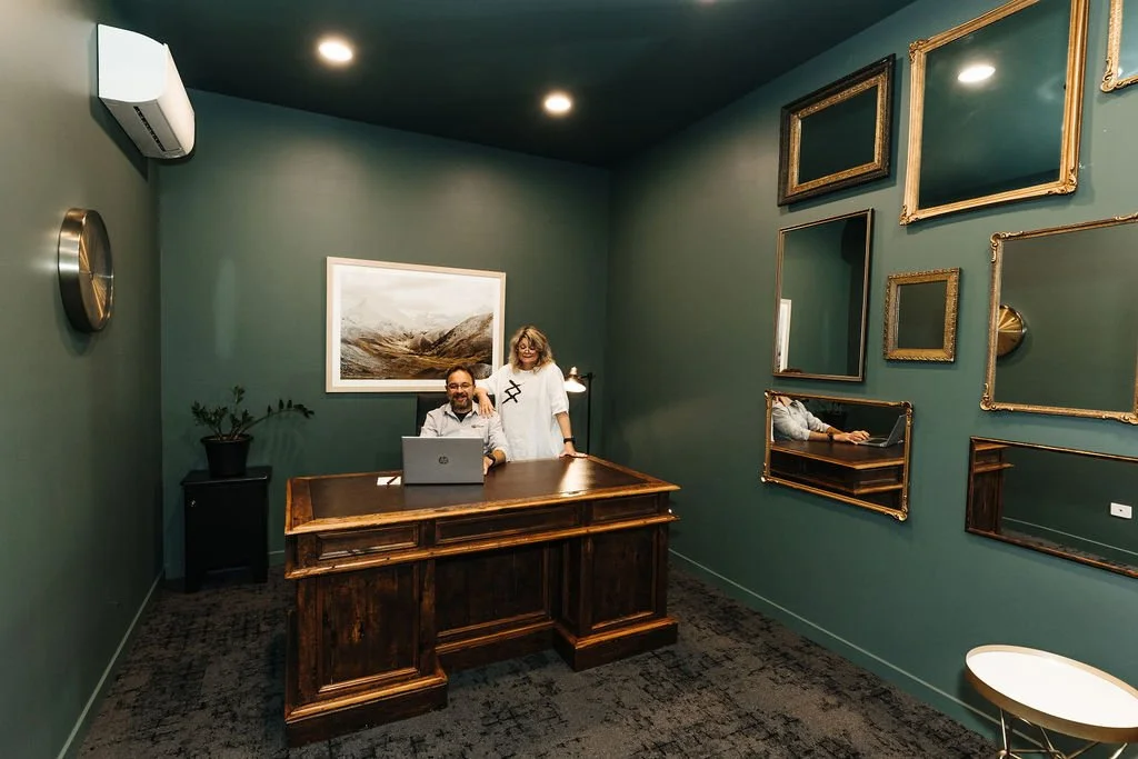 A man and woman in an office with green walls decorated with mirrors, a large landscape painting, a plant, and mirrors, sitting and standing behind a wooden desk, with a laptop open, in a cozy, well-lit space.