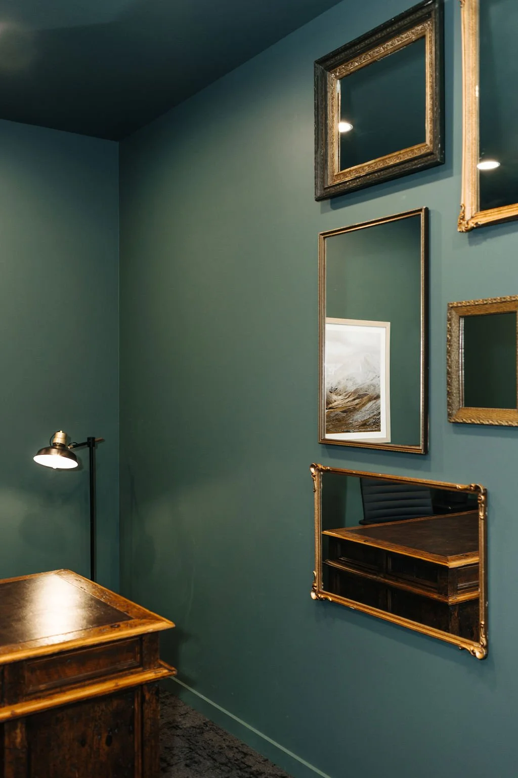 A corner of an office with dark green walls decorated with various mirrors and a framed landscape photograph. There is a vintage wooden desk in the foreground, and a black floor lamp with a metallic shade near the corner.