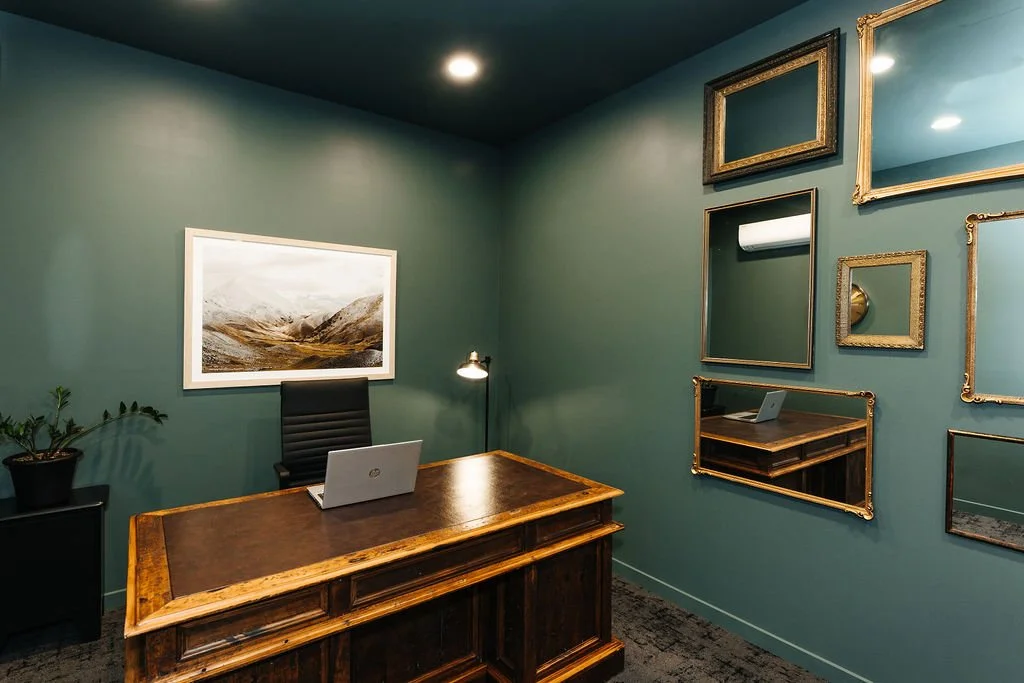 An office for lease at The Business Hive with a wooden desk, a black leather chair, a laptop, a potted plant, a large landscape painting on the wall, and several empty picture frames arranged decoratively on the teal wall.