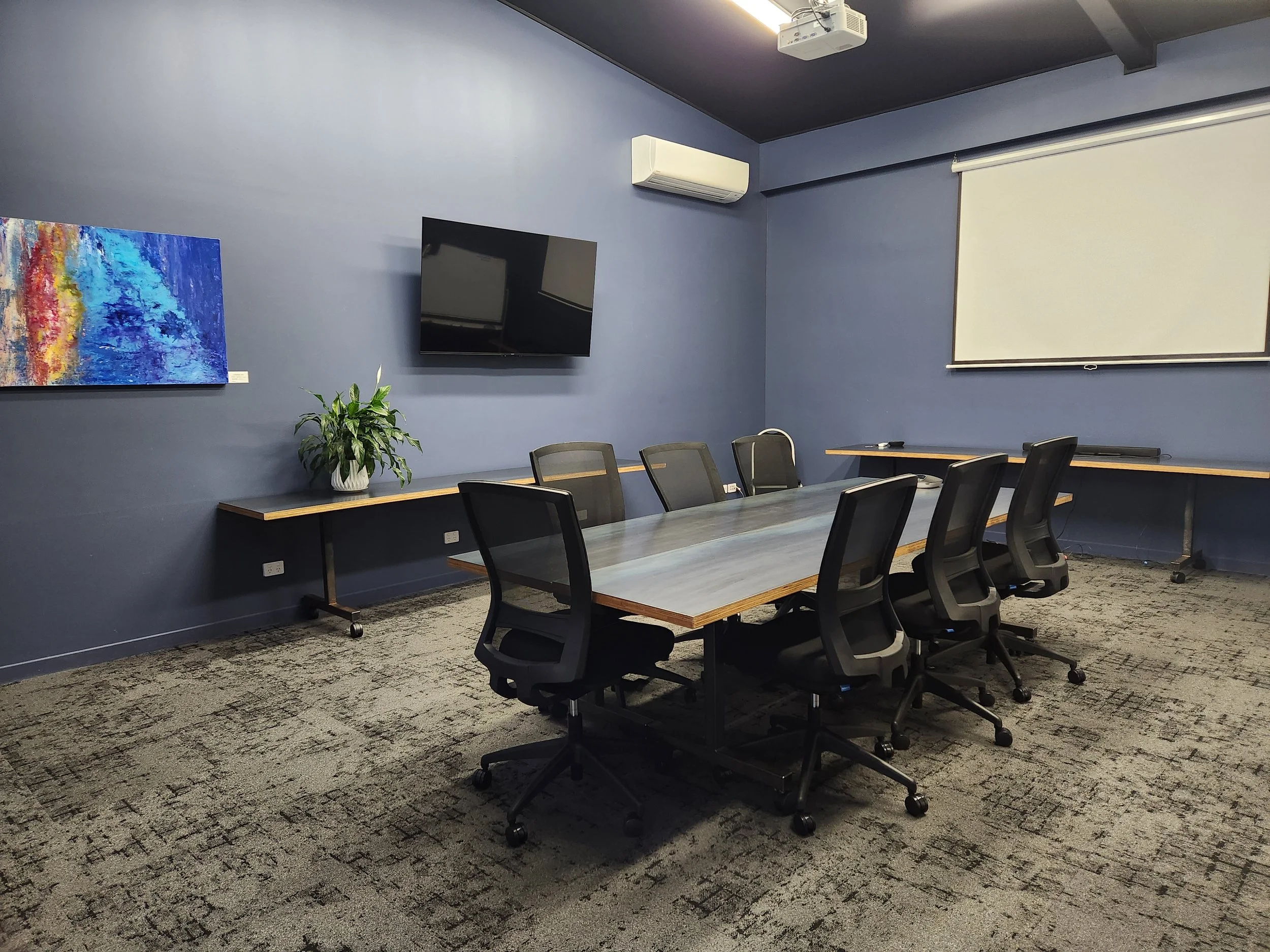 Meeting and training room in Oamaru with a large conferencing table with six desk chairs, blue walls and a white air conditioning unit and a large flat screen TV on the wall