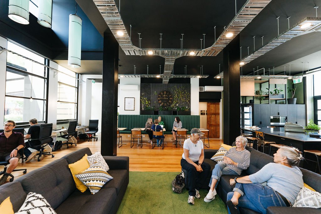 Open plan coworking space at The Business Hive in Oamaru with two couches, cafe style tables, desks and a large kitchen island. It has white walls, large windows and a black ceiling with people sitting and engaging in conversation