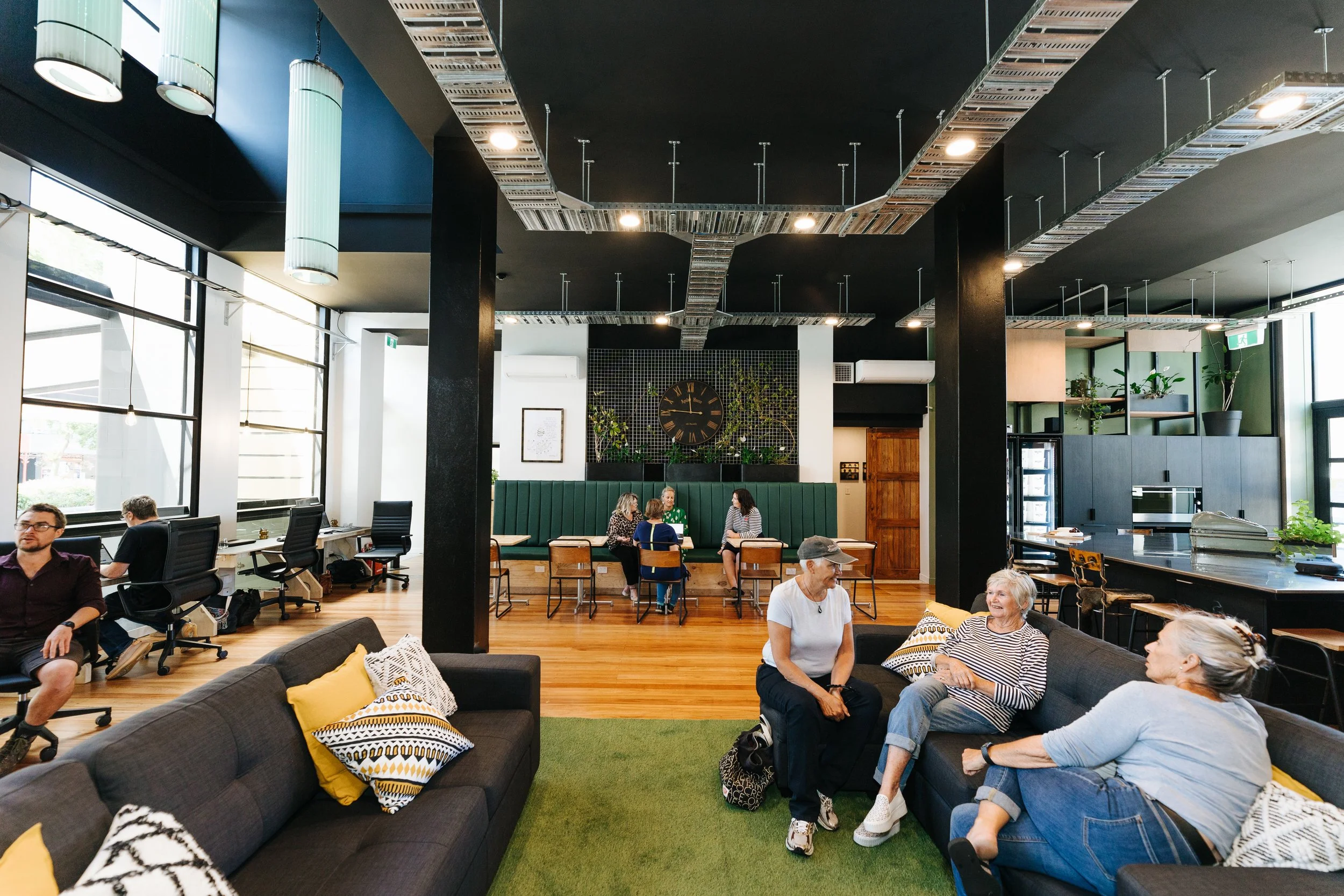 The Bank event and conference space in Oamaru with one group of three people sitting talking on a couch and another group of four talking around cafe style tables in a room with wooden flooring and a black ceiling