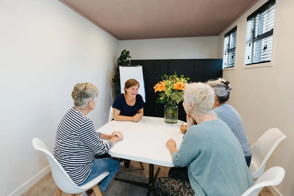 Meeting room in Oamaru with four women sitting around a white table with a vase of orange flowers talking and laughing with black lockers and a plant in the background.