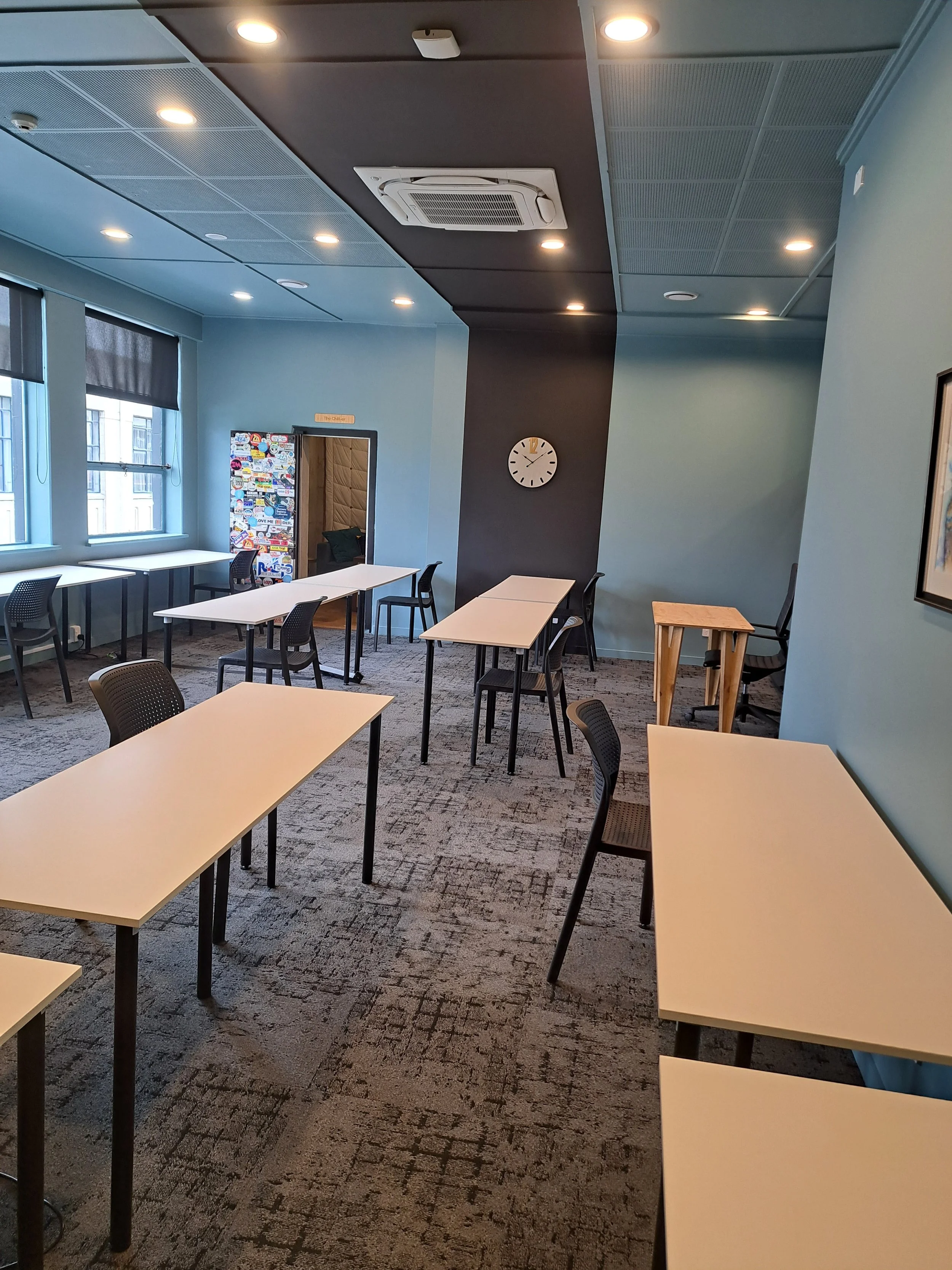 Empty training room with multiple white tables and black chairs, large windows with blinds, a clock on a dark accent wall, and a carpeted floor.