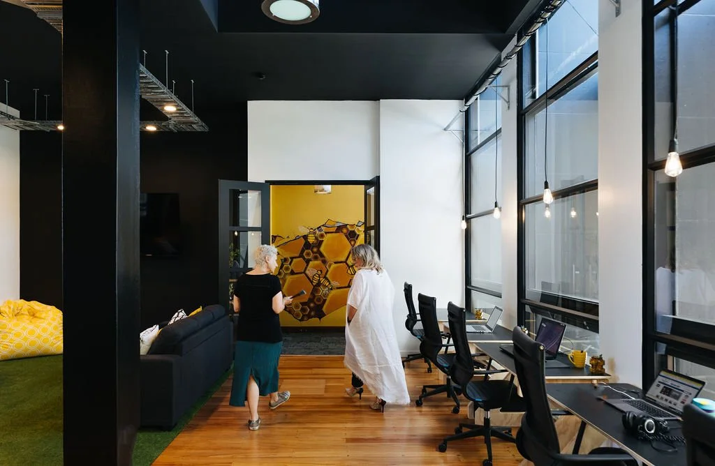 Two women talking while walking in a coworking space Oamaru featuring a wooden floor, black and white walls, black desks with black office chairs and large glass windows