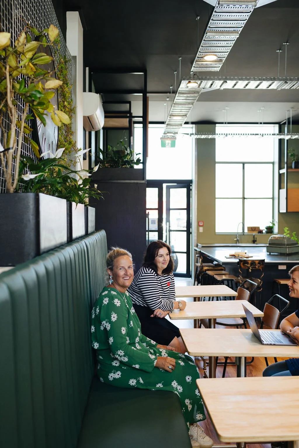 Three women engaging in conversation at cafe style tables at a coworking space in Oamaru with a long green seat and an open door and a large kitchen island in the background