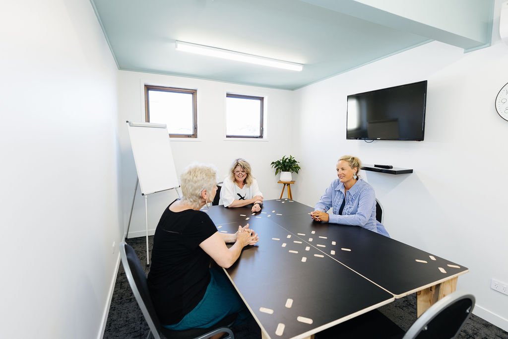 Bright meeting room in Oamaru with three women sitting around a black table in a space with white walls, a small whiteboard, a tv on the wall, two small square windows and a pot plant in the corner