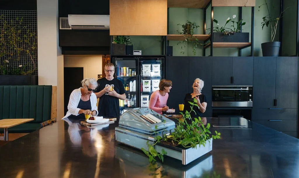 Communal kitchen with a large kitchen island at The Business Hive where four people laugh and engage in conversation while drinking coffee in front of a double glass door fridge