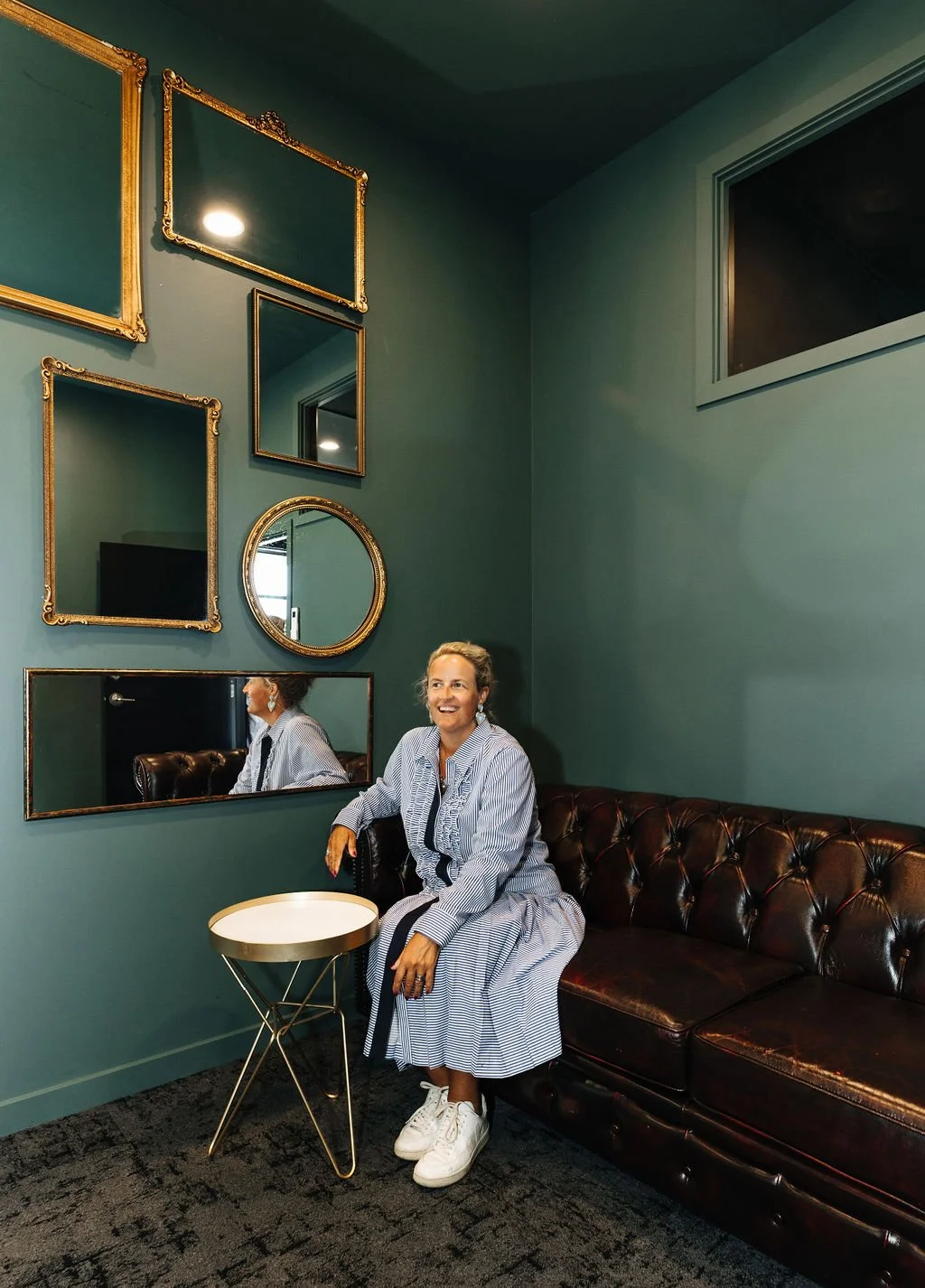 A woman sitting on a dark brown leather tufted sofa, smiling in The Mill, a private office at The Business Hive coworking space. It has green walls decorated with gold-framed mirrors and pictures.