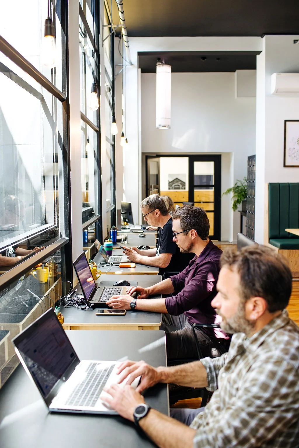 Coworking desks at The Business Hive with people sitting at black desks working on laptops in front of large glass windows in a white painted space with black lockers and double glass doors in the backgroun