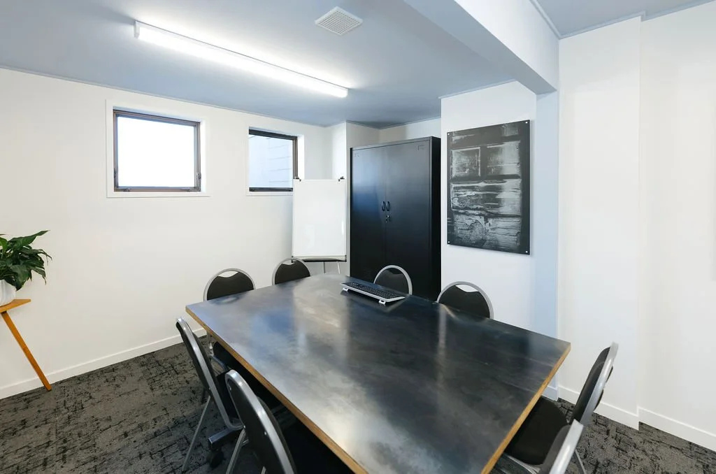 Bright meeting room in Oamaru featuring a black table with eight black chairs with metal rims, white walls and a whiteboard, a black cupboard a black and white art piece on the wall