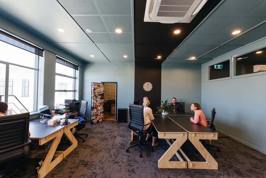 The Works private office space in Oamaru with black desks in a light blue room with a wide black stripe running up the wall and over the ceiling. Four people are having a conversation around the desks