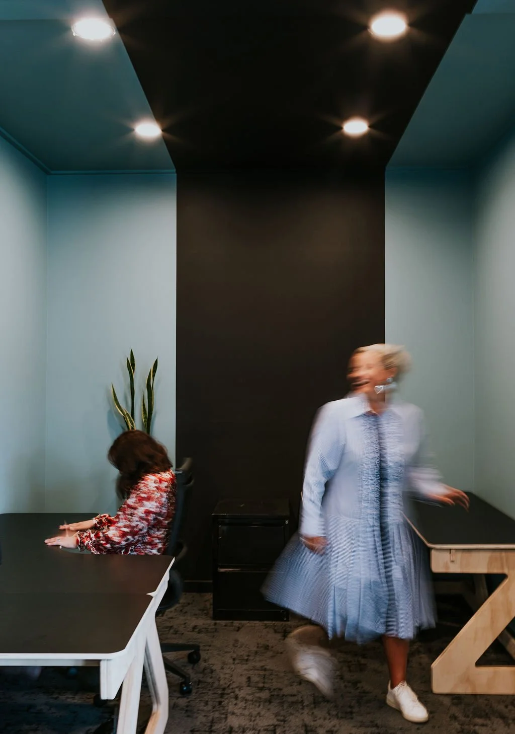 The Works private office Oamaru with one woman sitting at a black desk and another walking away in a blue room with a wide black stripe running up the wall and onto the ceiling