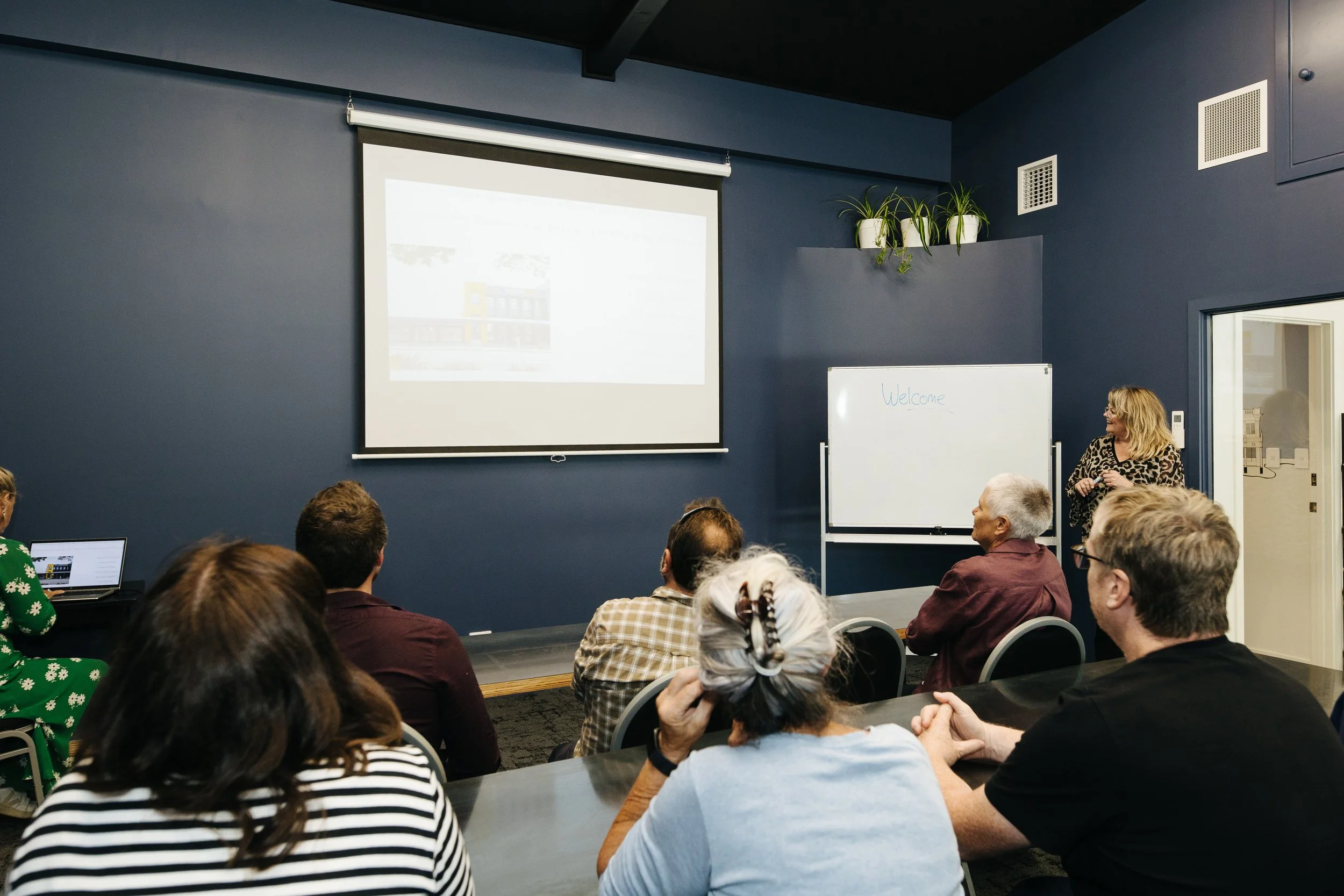 six people in a meeting room sitting at long tables and looking at a white projection screen which shows the front of The Business Hive coworking space, hung up against a blue wall