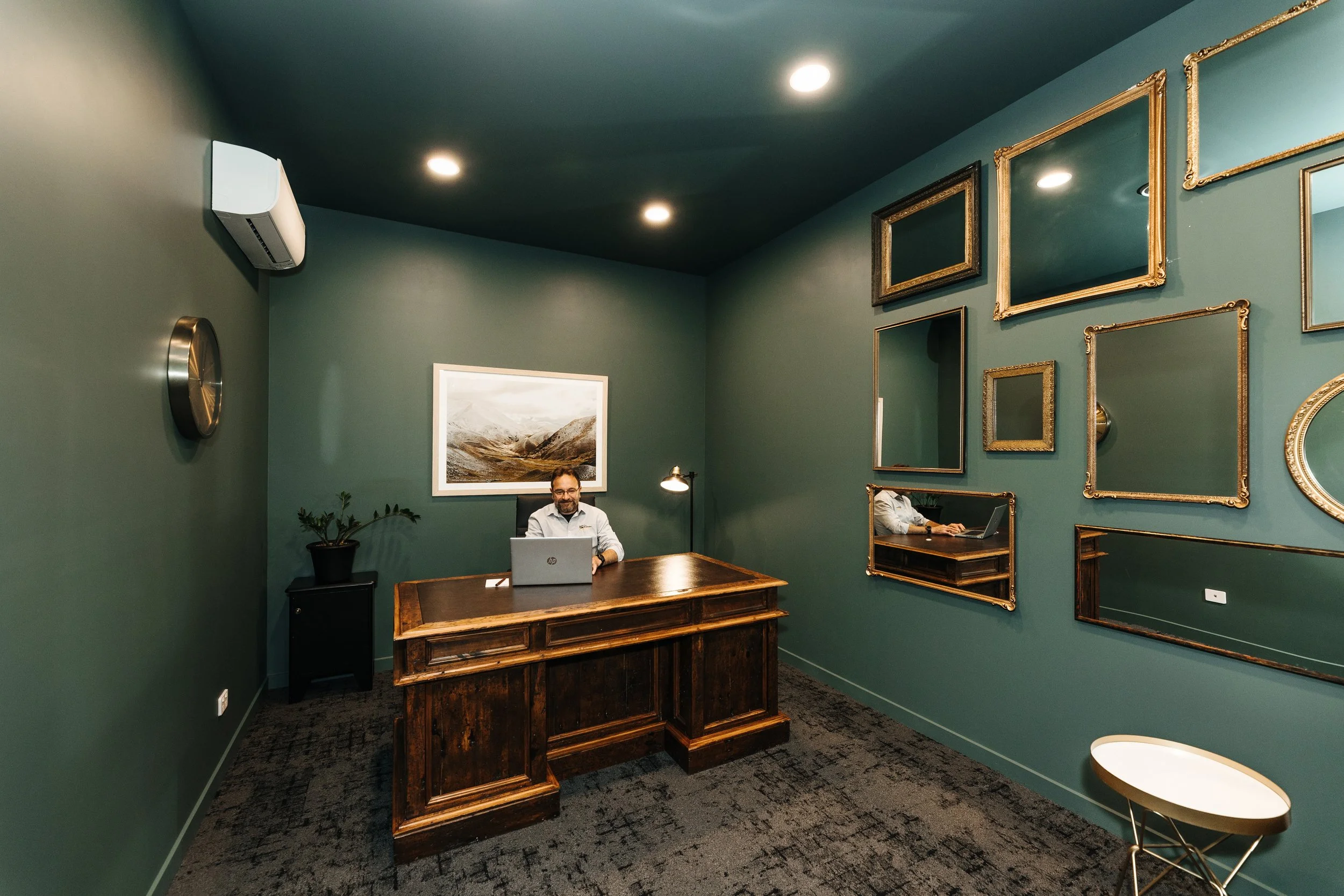 The Mill private office Oamaru where a man sits in front of a laptop at a large wooden desk in a dark green room lit by spotlights with mirrors on the wall, a framed picture behind him and a clock and air conditioning unit on the wall