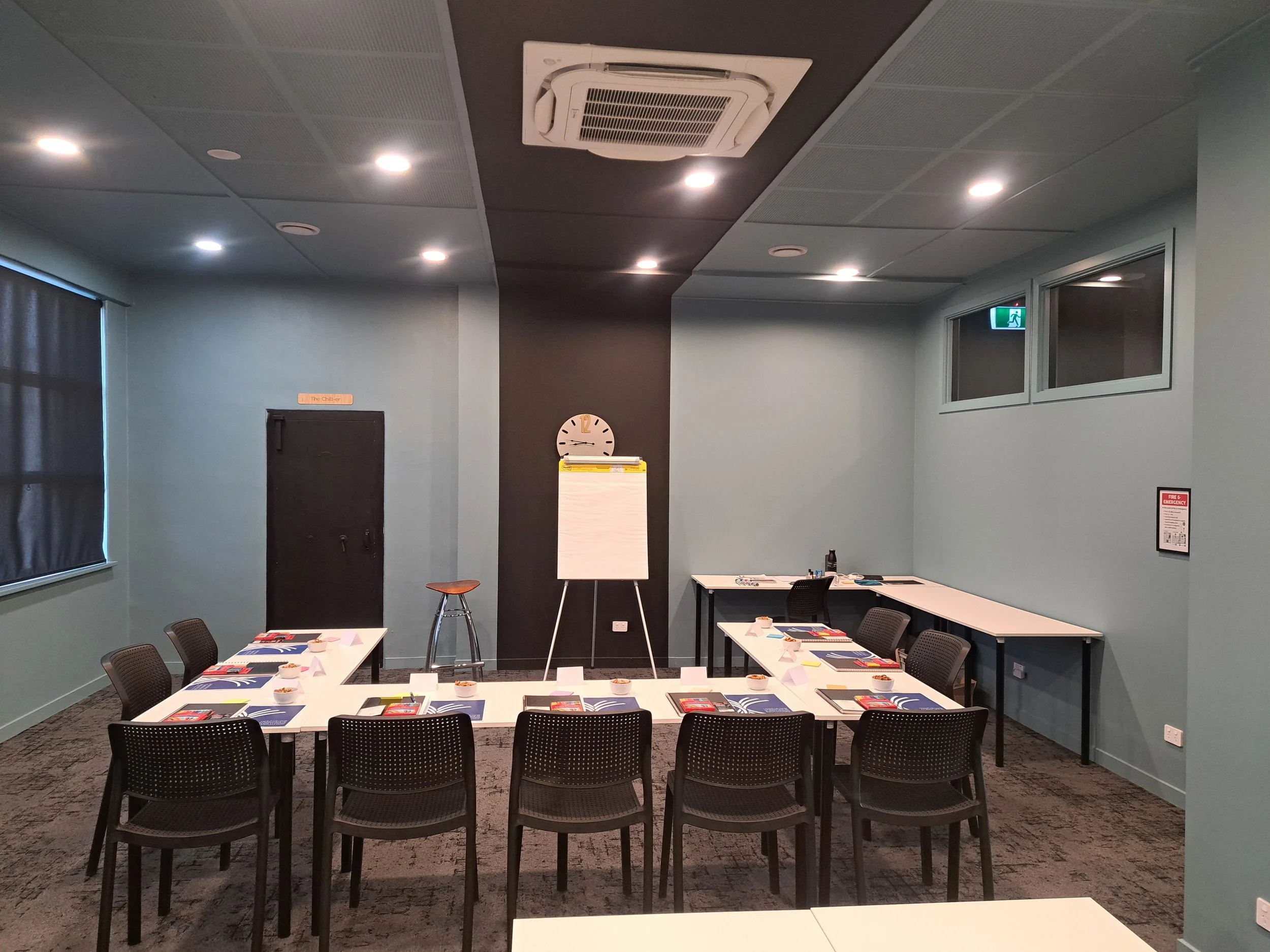 Empty training room with U-shaped table setup, chairs, notepads, and pens, with a whiteboard, clock, and a stool at the front, and windows with curtains on the side.