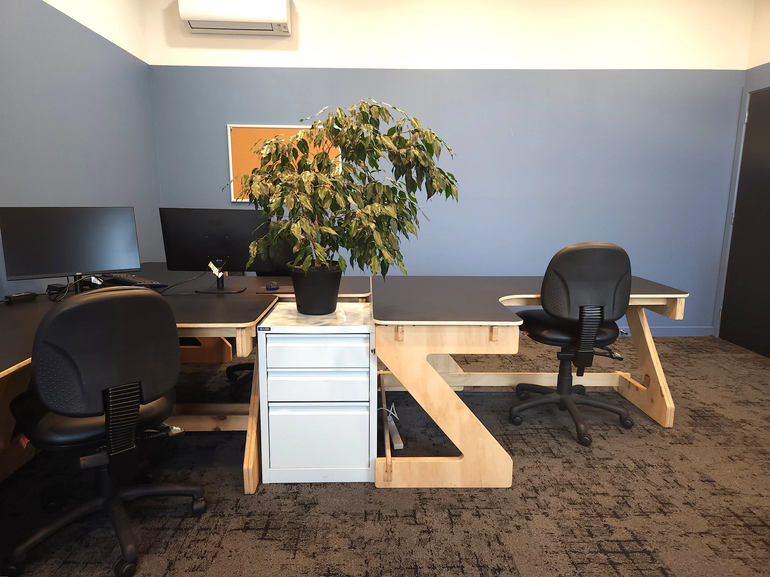 Office space with two black chairs, two computer monitors, a desk with a large potted plant, a white filing cabinet, and a blue wall with a corkboard. An air conditioning unit is mounted on the wall.