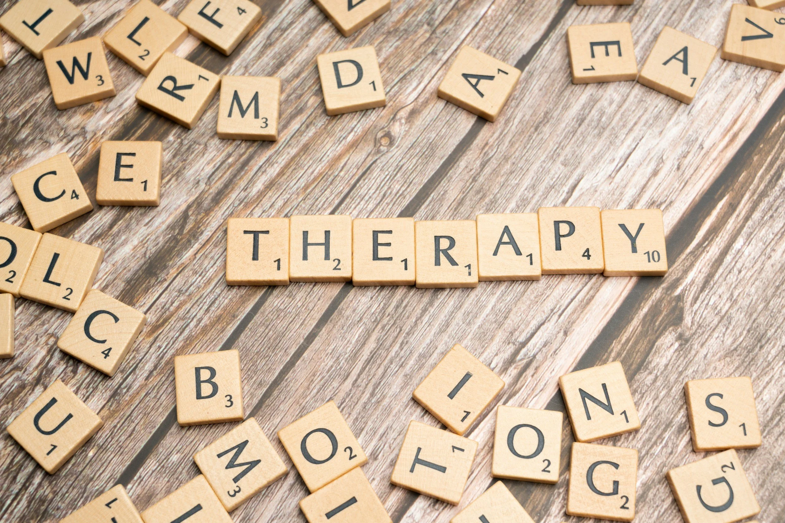 Scrabble tiles spell out the word 'THERAPY' on a wooden surface, with scattered letters and tiles around.