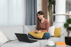 Woman sitting on a sofa with a laptop, holding a yellow pillow, in a living room with plants and decor.