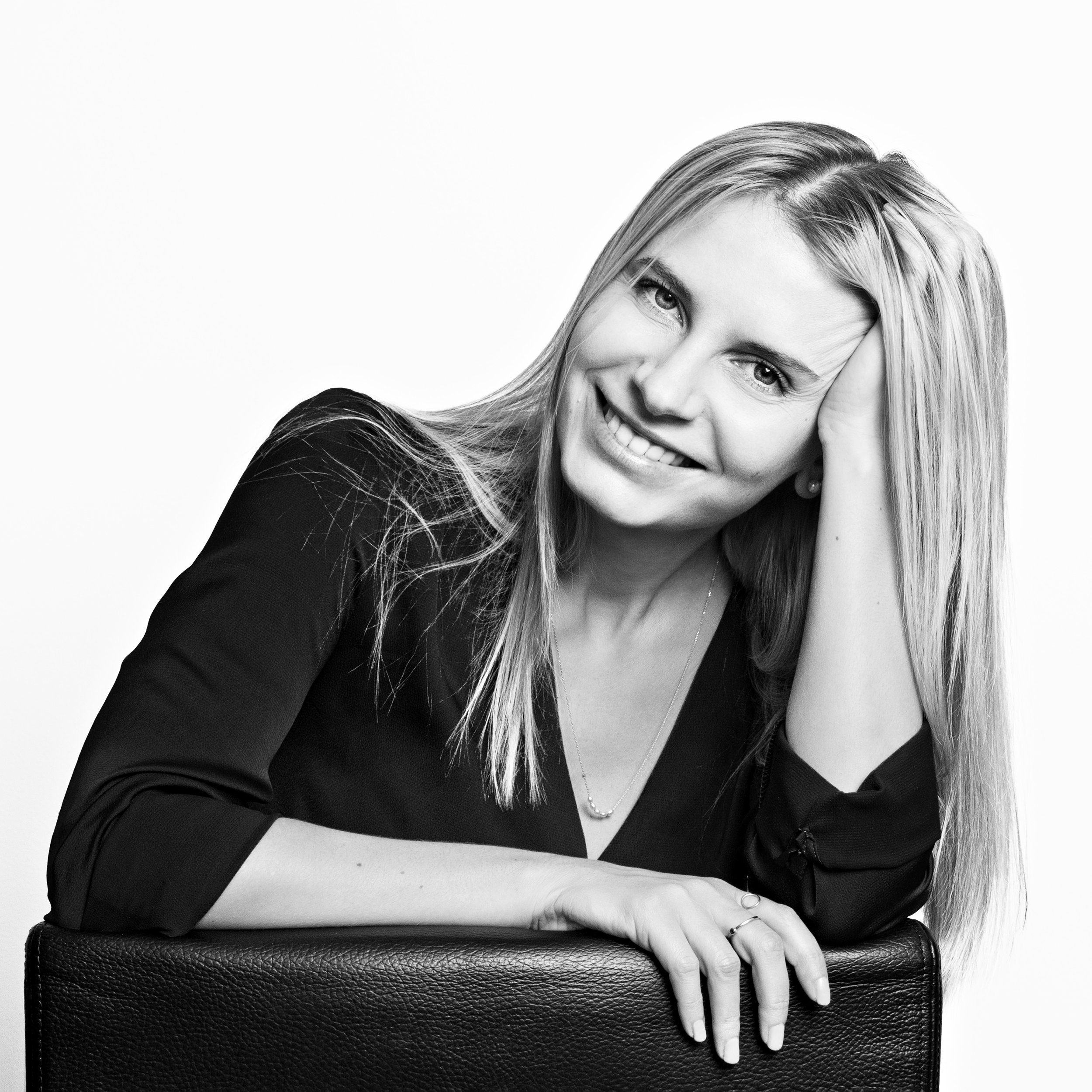 Black and white portrait of a smiling woman with long hair, leaning on a textured surface, wearing a dark top and a necklace.