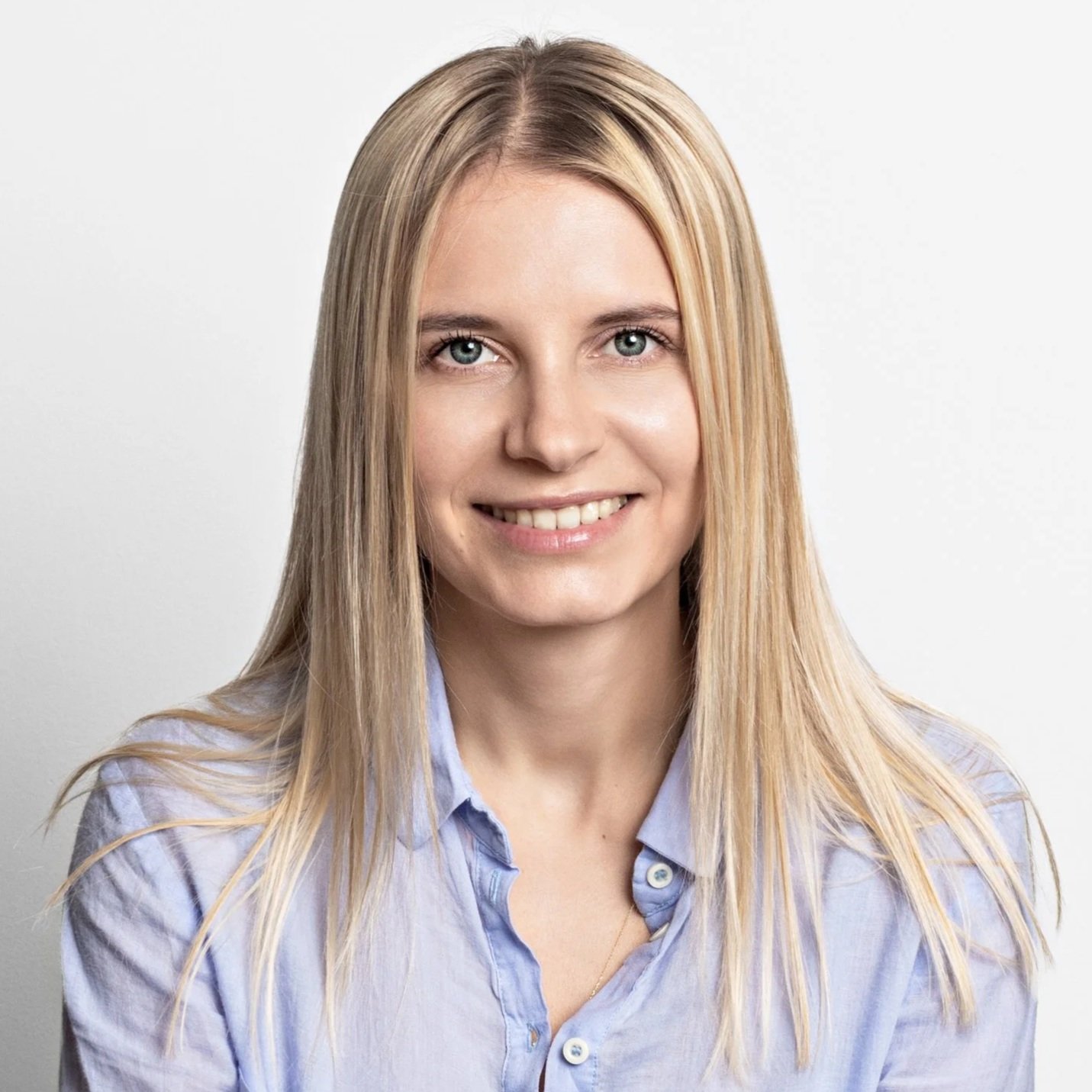 A young woman with long blonde hair and blue eyes, smiling, wearing a light blue button-up shirt, against a plain white background.
